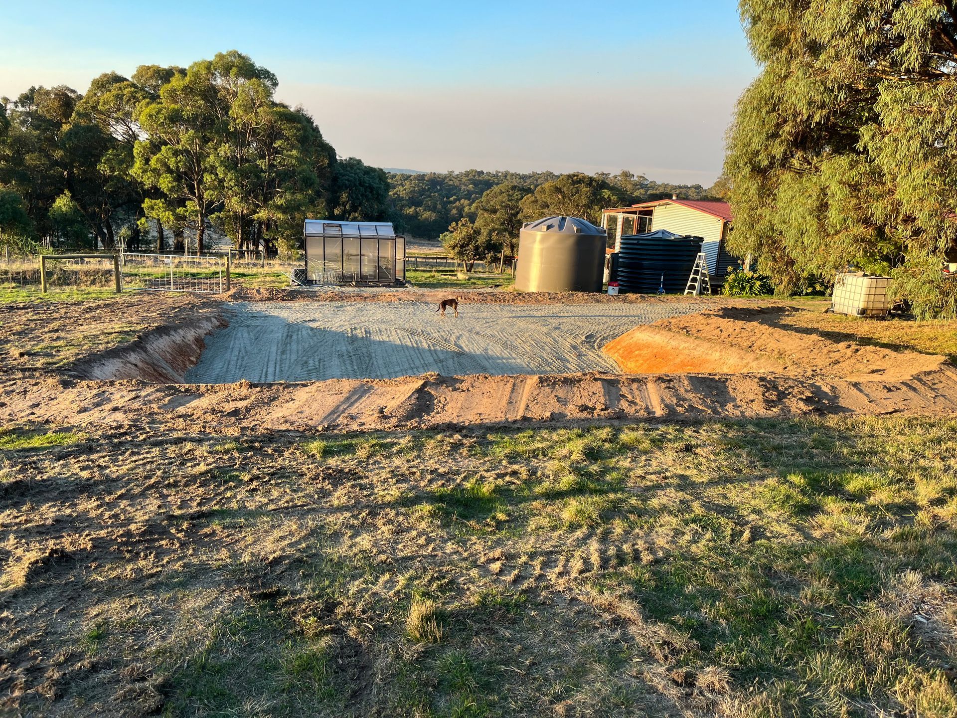 Construction site with excavated area, gravel, water tank, and trees under a blue sky — Beaufort & District Earthworks in Beaufort, VIC