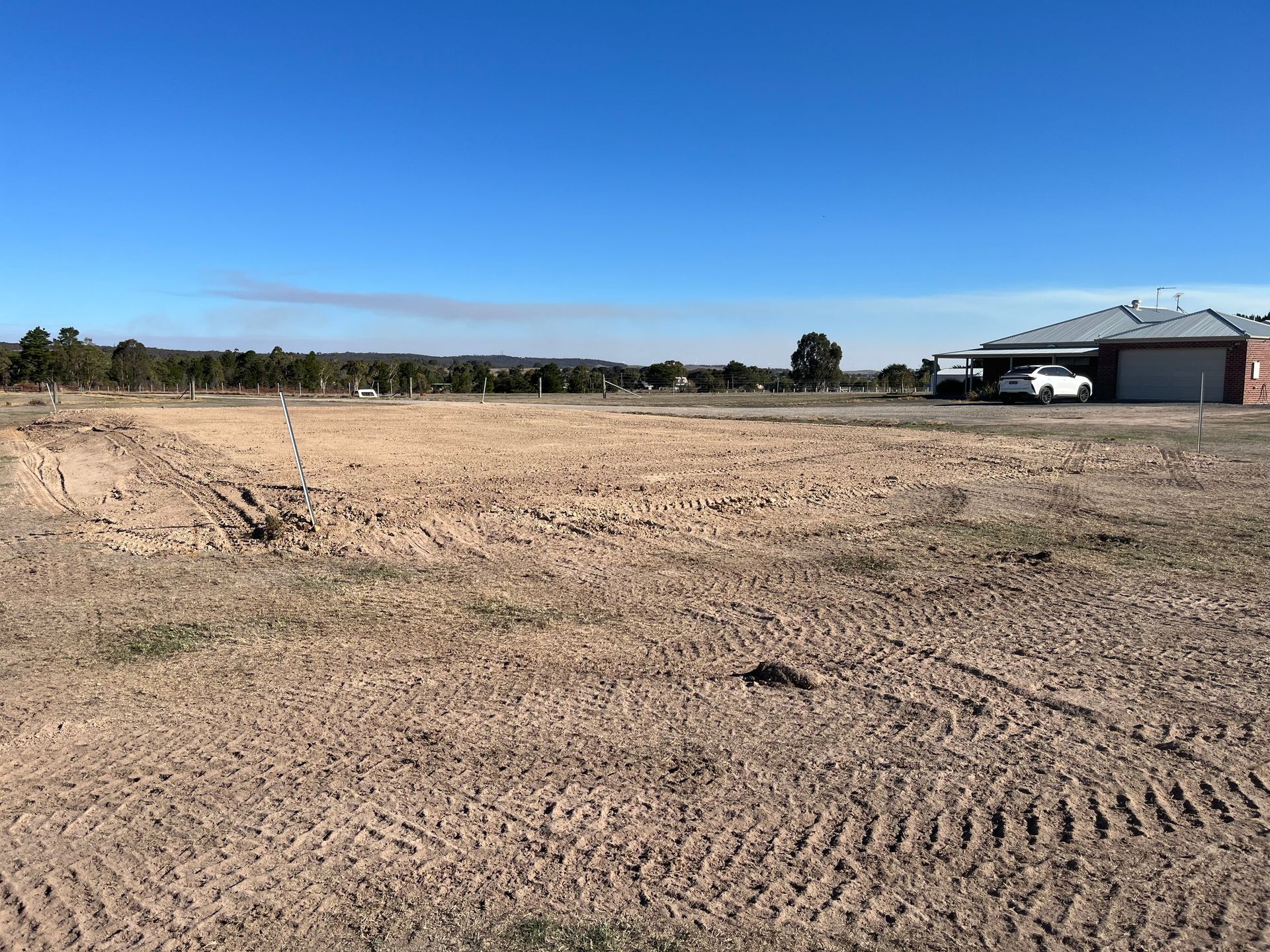 Empty dirt lot with tire tracks and a house with a car parked in the driveway under a bright blue sky — Beaufort & District Earthworks in Beaufort, VIC