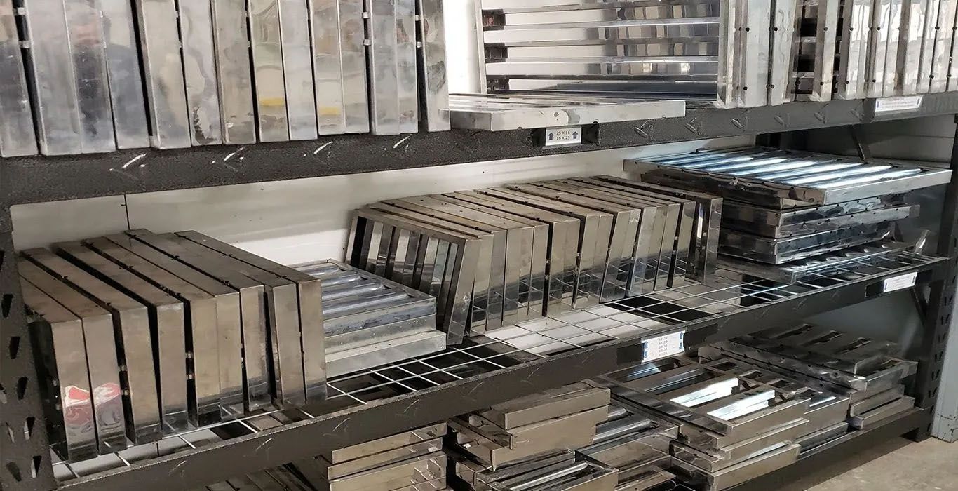 Shelves holding various metal parts; stacks, bars, and frames. Industrial setting, gray tones.