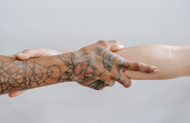 Tattooed hand clasps a lighter-skinned arm, close-up, against a plain background.