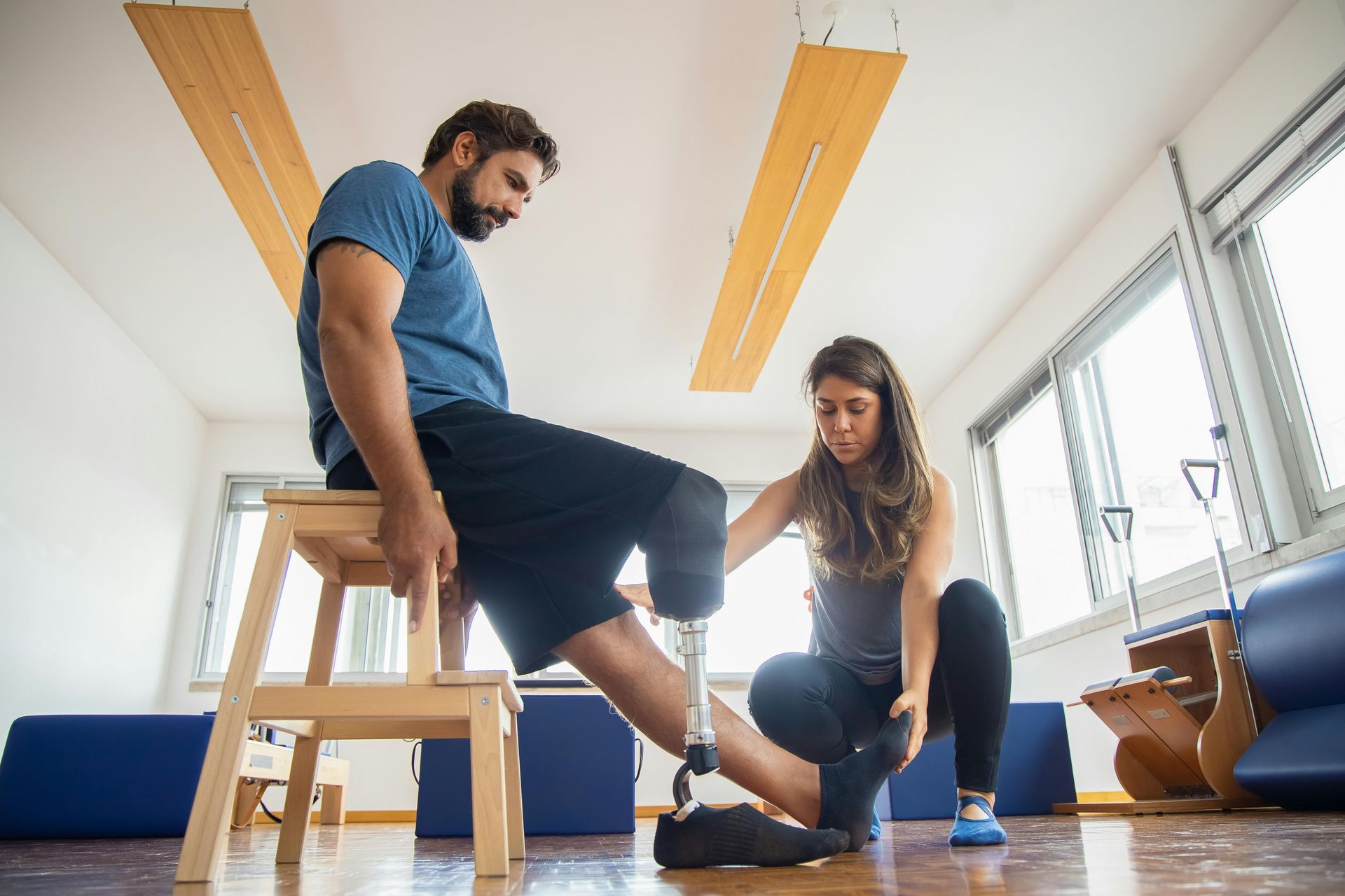 Man with prosthetic leg in therapy, therapist helping. Bright room, beige and blue accents.
