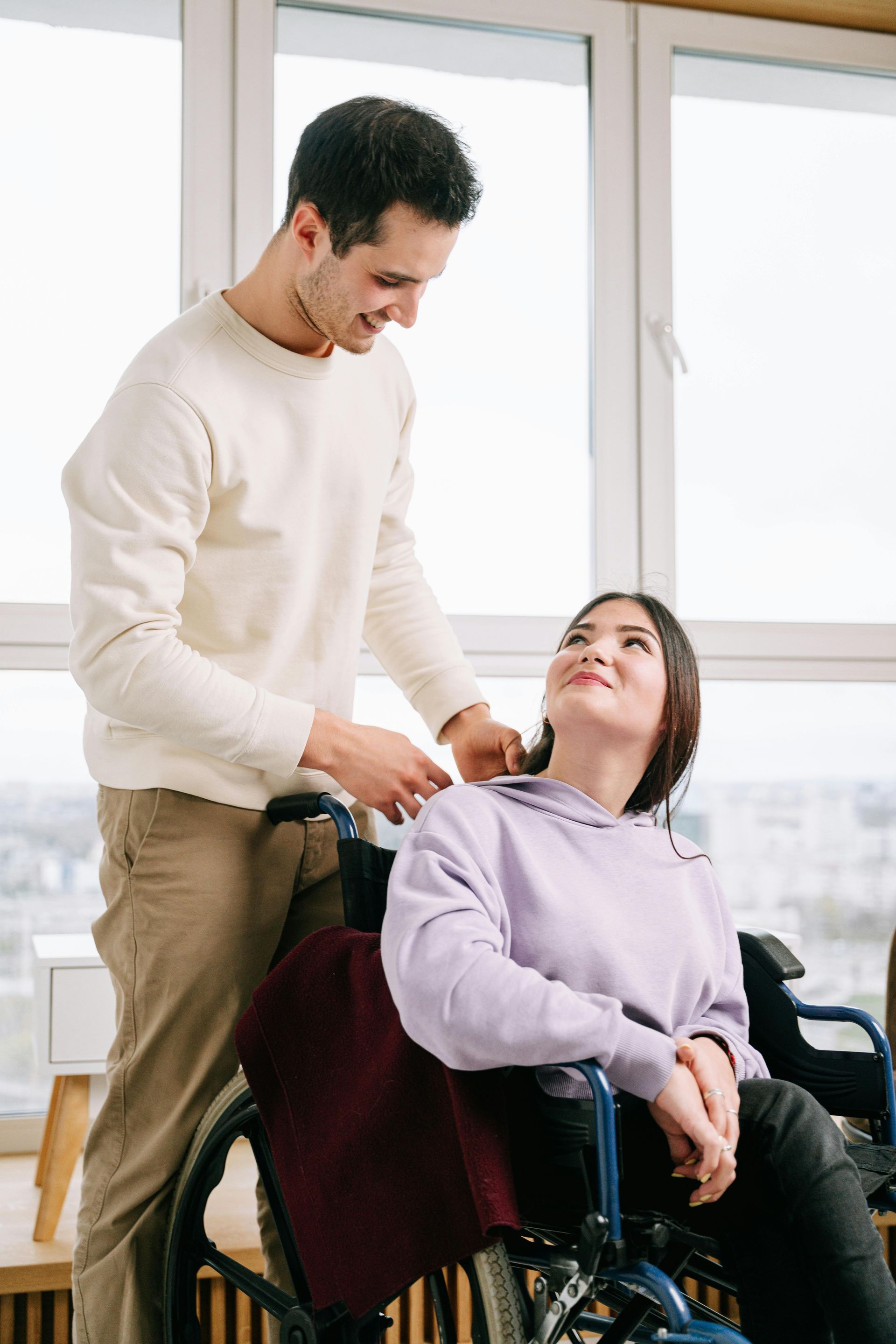 Man helping woman in wheelchair; smiling; indoors, near window.