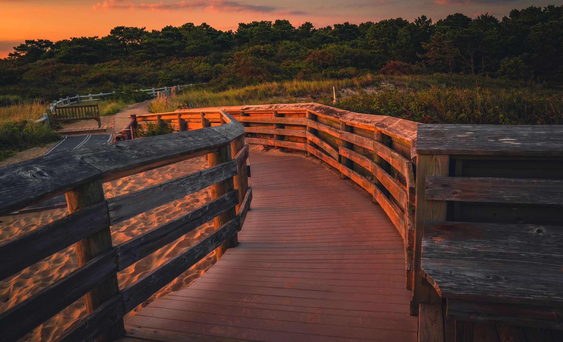 Wooden boardwalk with railings winding through a marsh at sunset, lit by warm orange light.