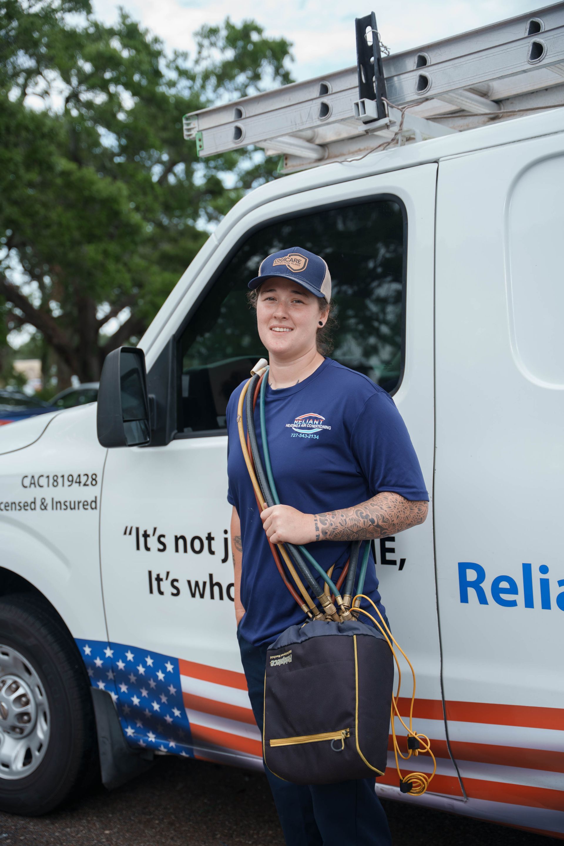 A Reliant Heating & Air Conditioning technician is standing in front of a van holding a bag.