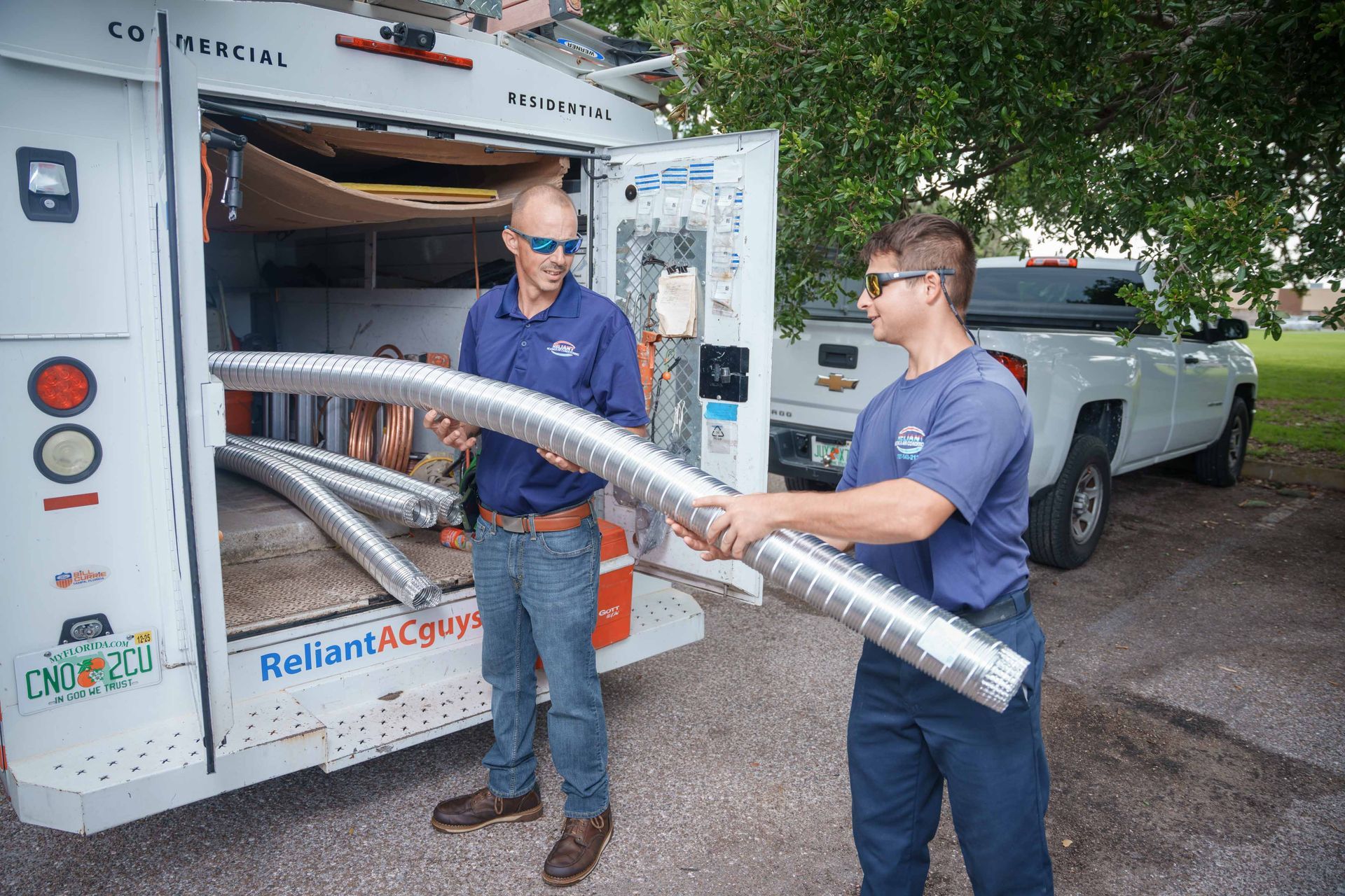 Two Reliant Heating & Air Conditioning technician are holding large pipes in front of a truck.