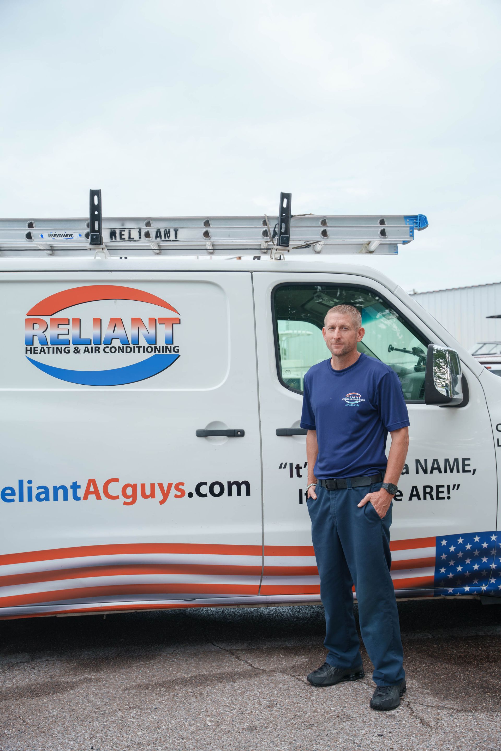 A Reliant Heating & Air Conditioning technician in a blue shirt is standing in front of a white van.
