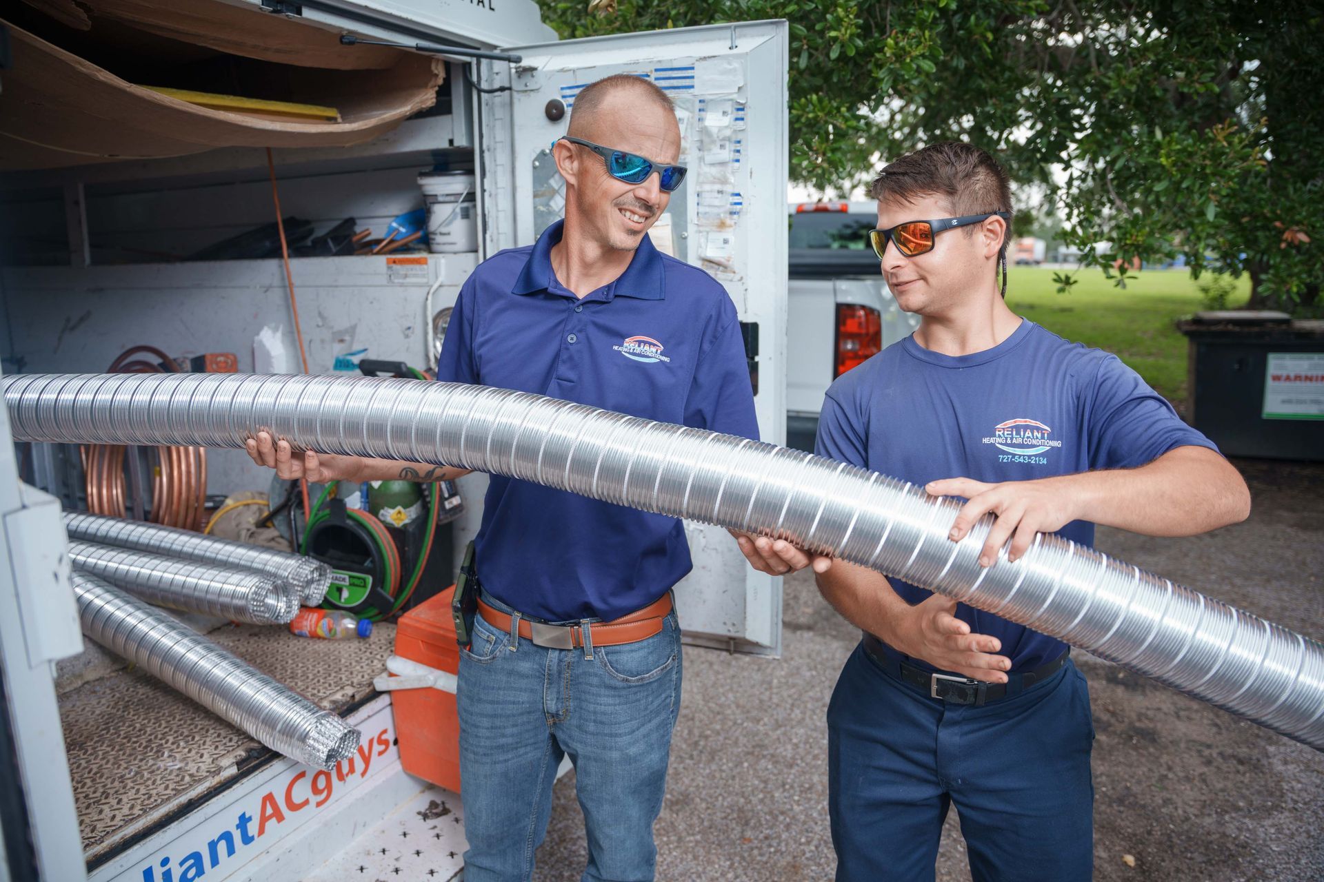 Two Reliant Heating & Air Conditioning technician are standing next to each other holding a large metal pipe.
