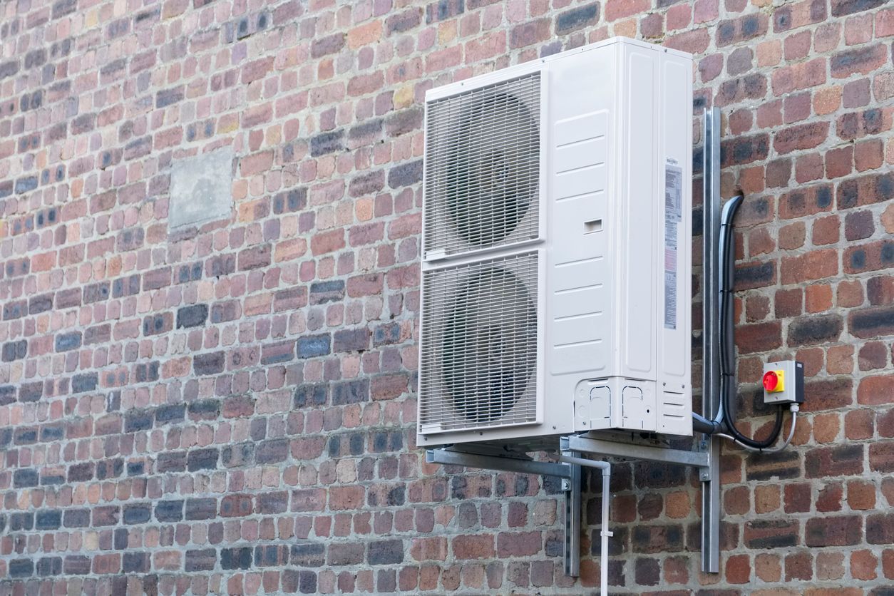 A white air conditioner is mounted on the side of a brick building Installed by Reliant Heating & Air Conditioning.