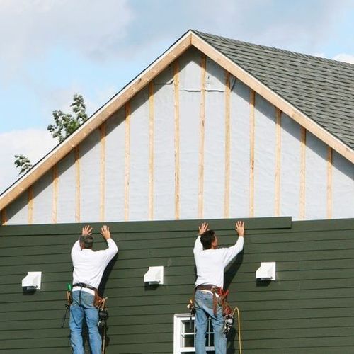 Two construction workers installing siding on a house exterior. Green siding, wood frame, blue sky.