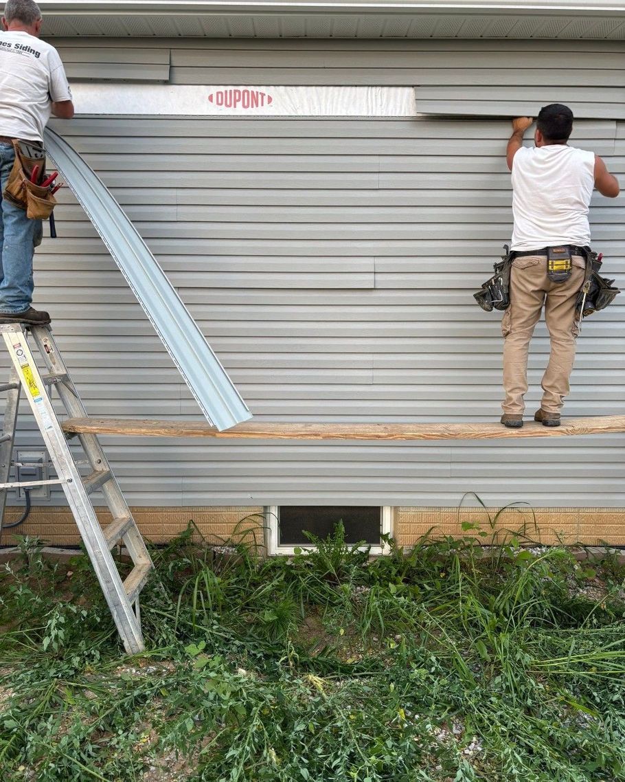 Two workers installing siding on a house, using a ladder and a plank as a scaffold.