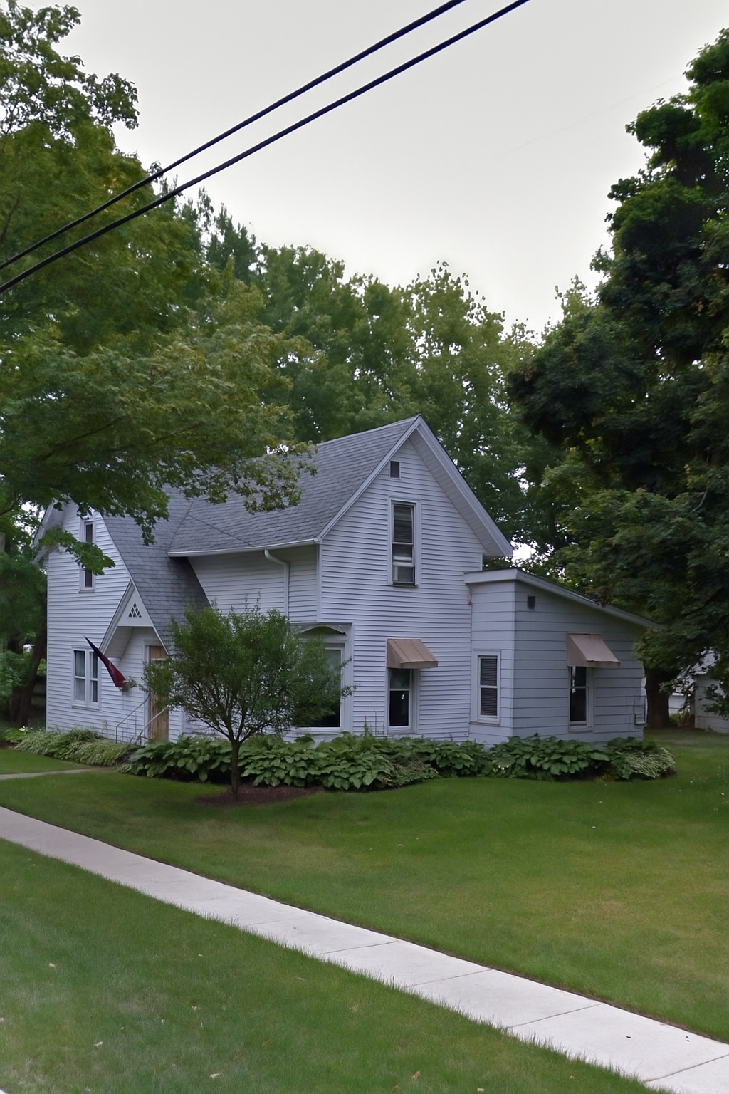 Light blue two-story house with a gabled roof, surrounded by green trees and a sidewalk in front.