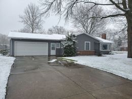 Gray house covered in snow with a driveway, garage, and bare trees in the background.