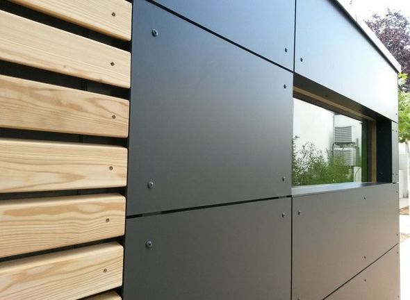 Exterior wall with horizontal wooden slats and black metal panels. A rectangular window reflects greenery.