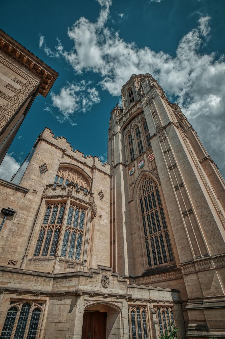 Stone university building with a tall tower, against a partly cloudy blue sky.