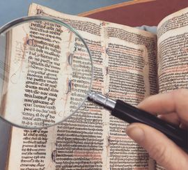 Hand holding magnifying glass over an open, old book with handwritten text, possibly a historical manuscript.