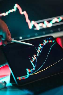 A hand holding a stylus points at a stock chart on a tablet, with a second chart on a monitor.