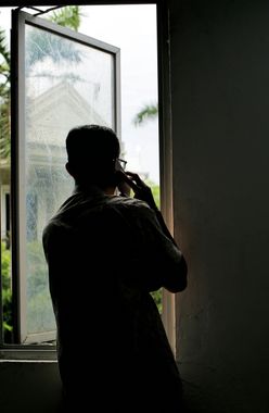Silhouette of a person talking on the phone near an open, rain-streaked window; outside, a glimpse of green foliage.