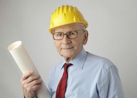Elderly architect wearing a yellow hard hat, glasses, and red tie, holding blueprints.