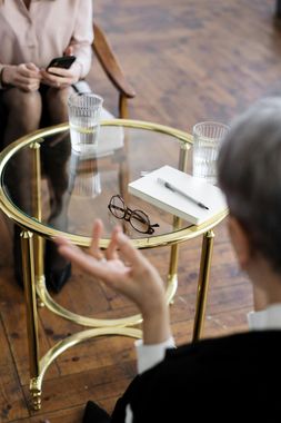 Two people in therapy session, one gesturing with hand, notes and glasses on table.