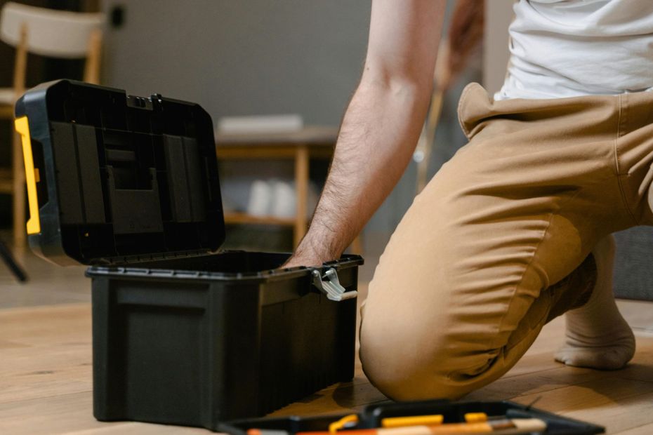 Person kneeling, opening a black toolbox on a wooden floor; home interior.