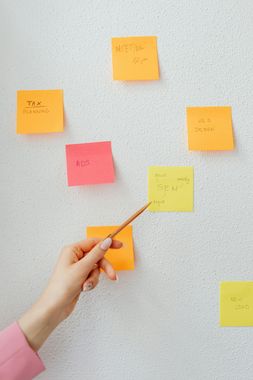 Hand pointing at a yellow sticky note on a white wall covered in similar notes of various colors.