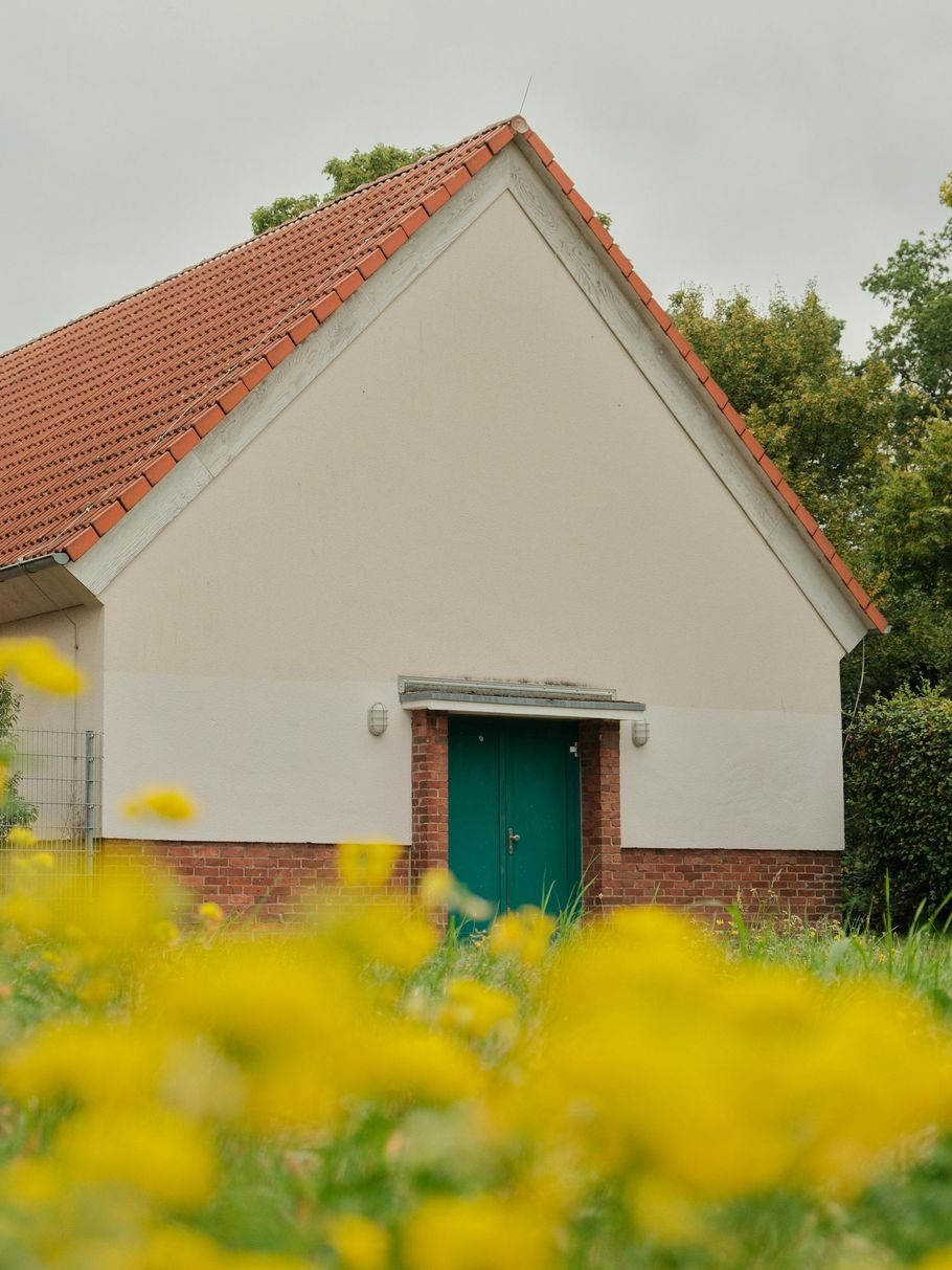White building with red tile roof and green door, surrounded by yellow flowers.