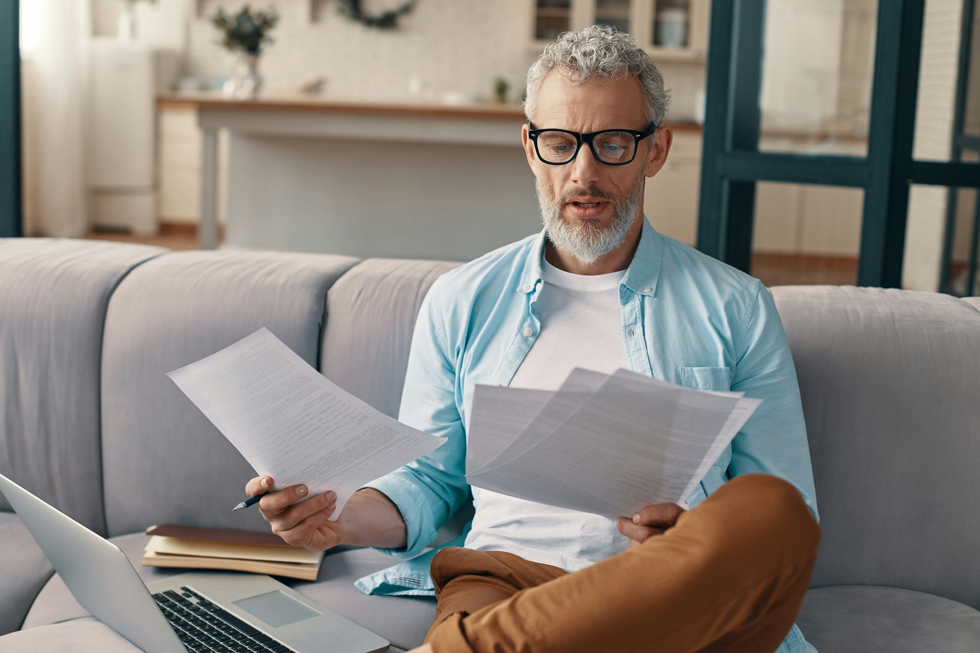 Man on a couch reviewing papers with a laptop, wearing glasses and a light blue shirt.