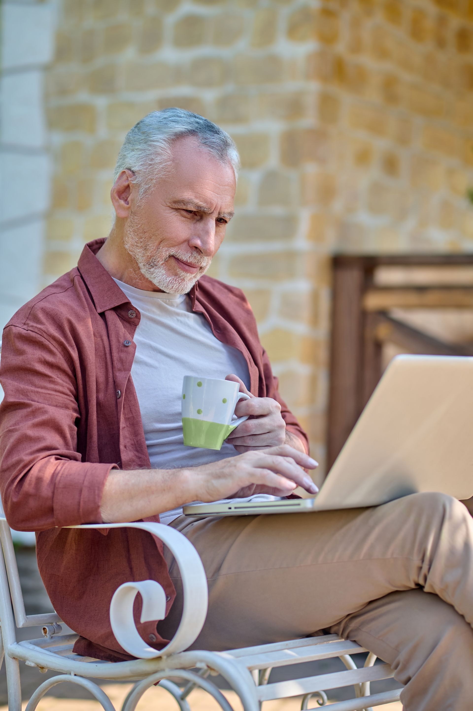 Man with gray hair, using a laptop, holding mug, sitting outside.