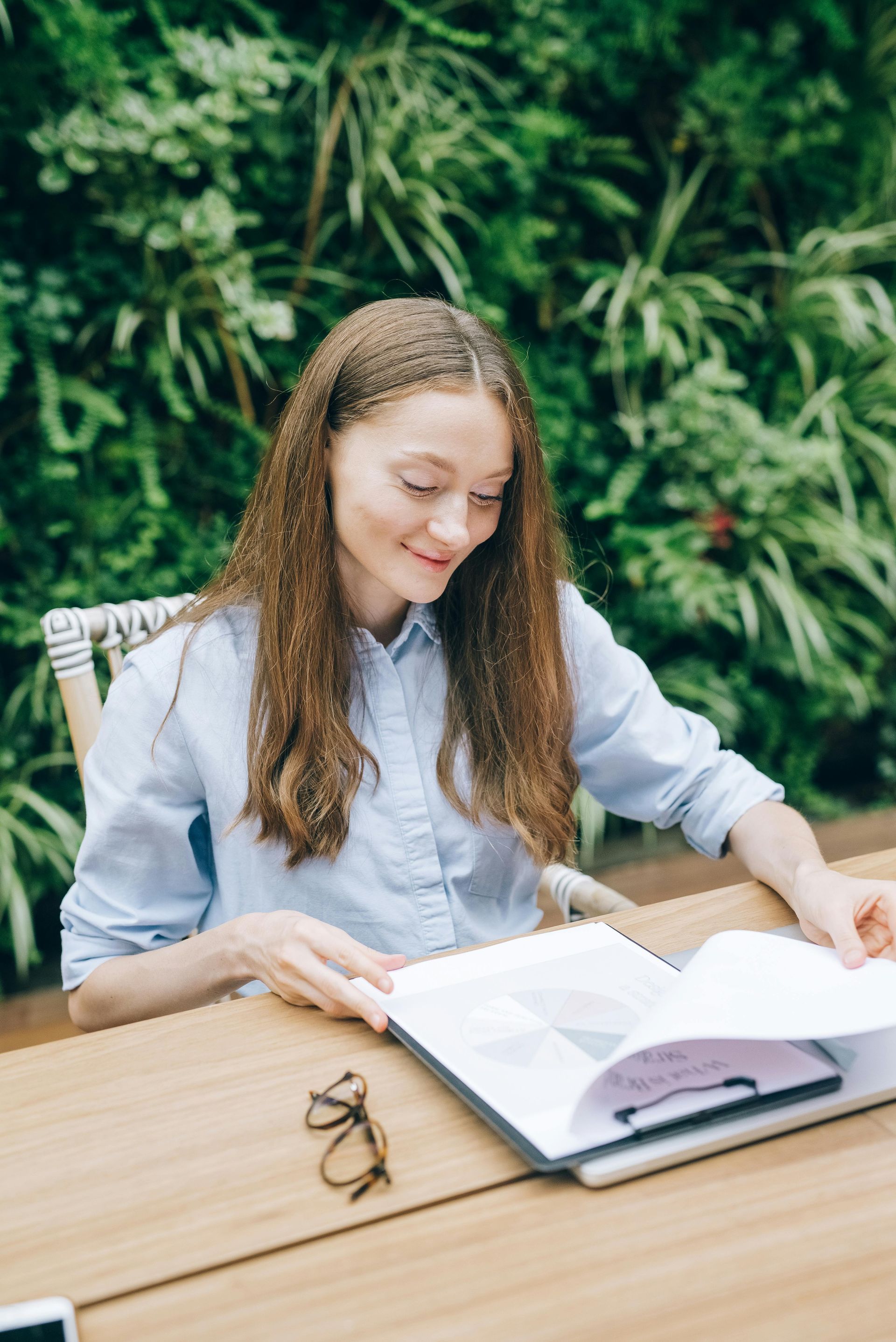 Woman reading book at table, smiling, outdoors, greenery background.