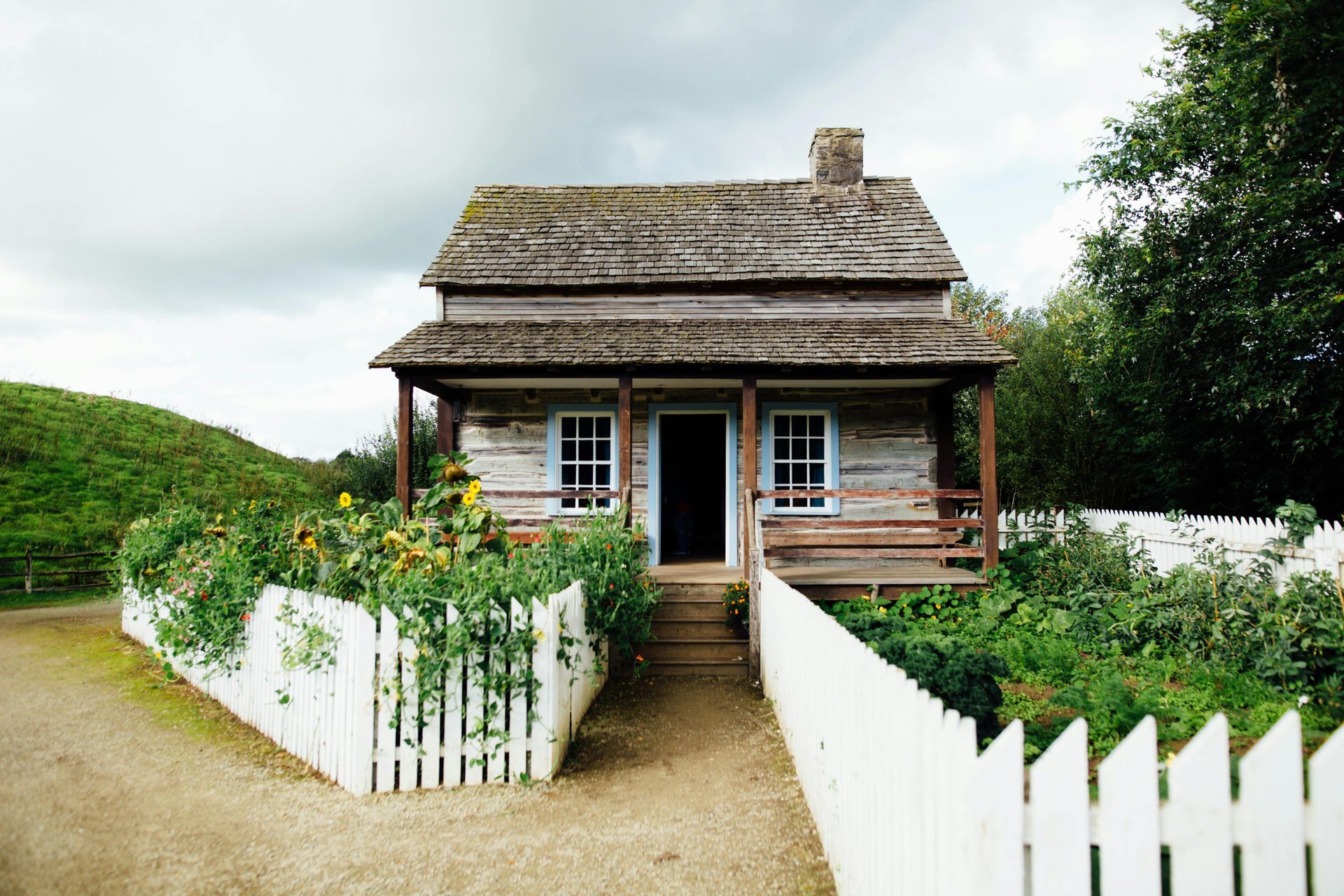 Small wooden house with porch, white picket fence, and garden. Cloudy sky.