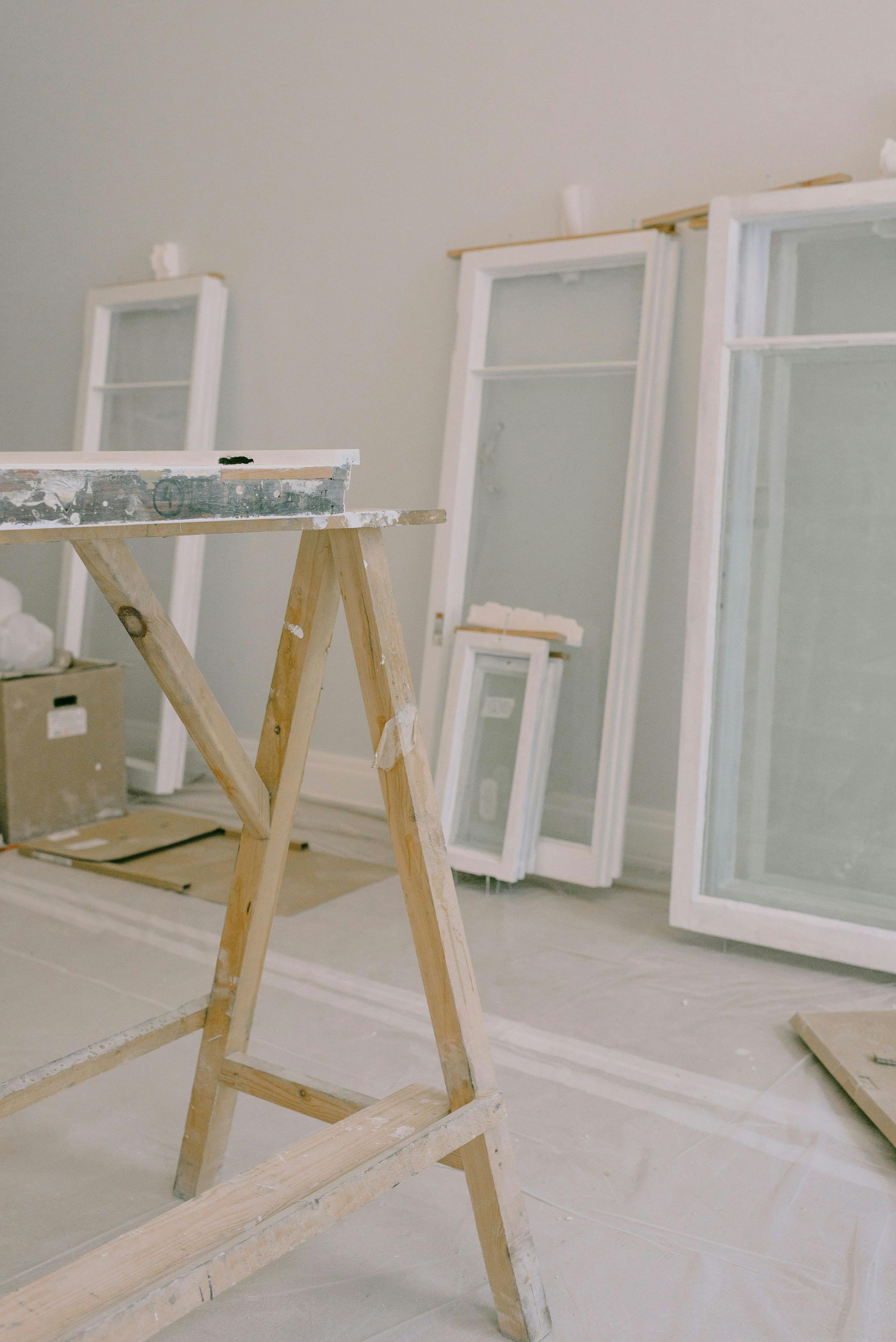 Construction scene: wooden sawhorse, multiple white window frames leaning against a wall, and painter's tarp on the floor.