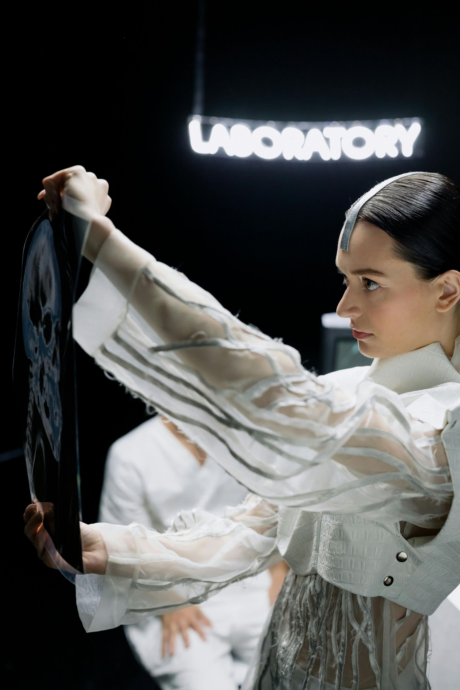 Mulher de jaleco branco examinando raio-X em um laboratório escuro; placa com a palavra 