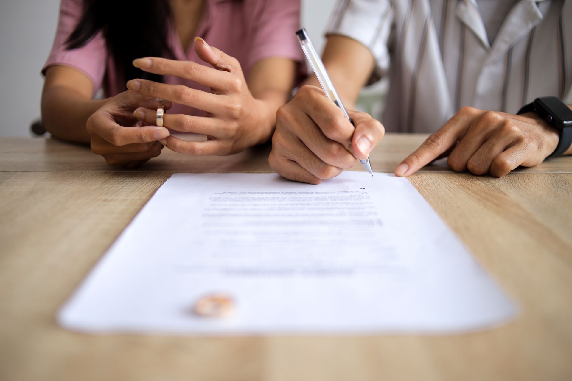 Couple signing a document, one holding wedding ring, wooden table.