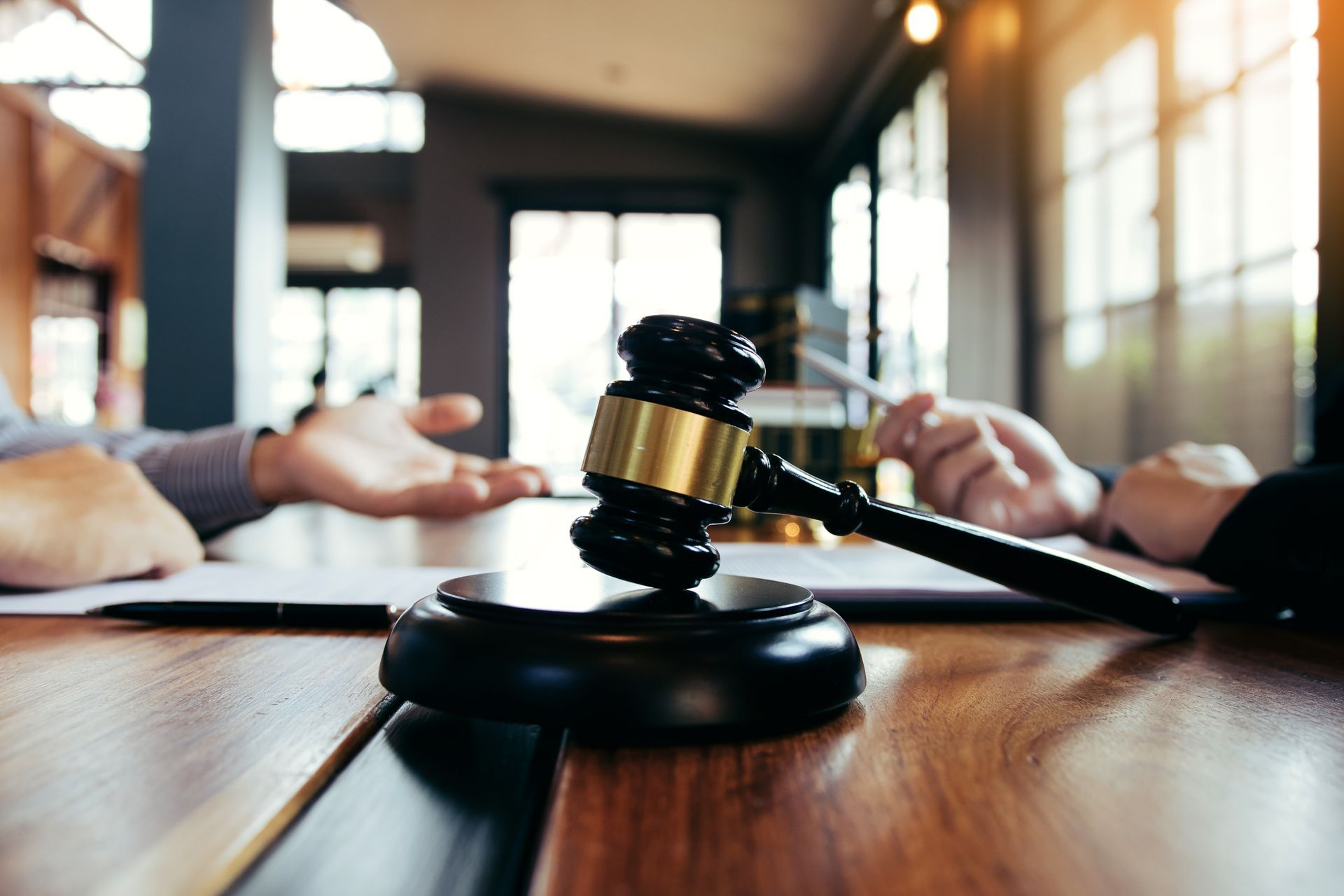 Judge's gavel on a wooden desk with two people in the background, possibly in a courtroom or legal setting.