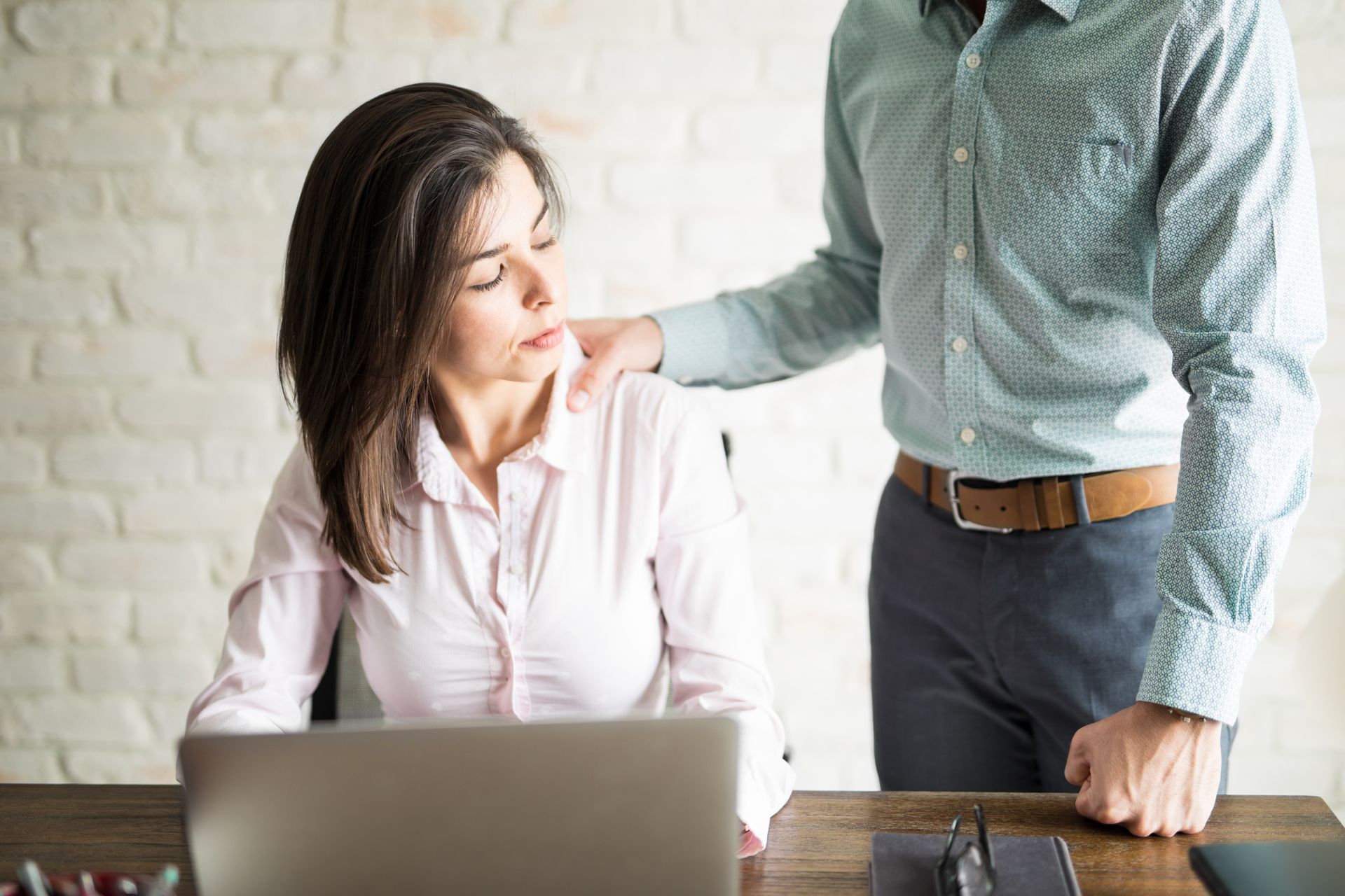 Woman at a desk looks uncomfortable as a man's hand rests on her shoulder.