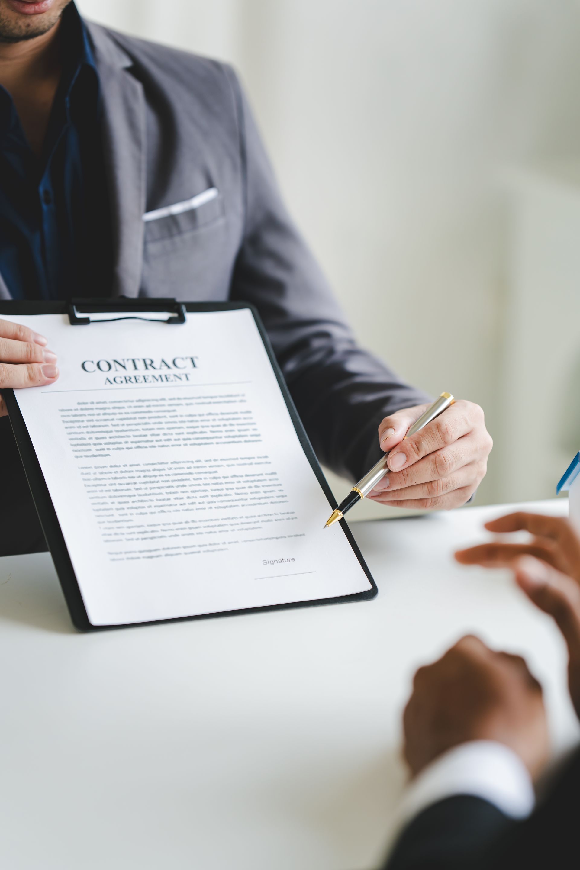 Person in suit pointing pen at a contract on a clipboard, sitting at a table.