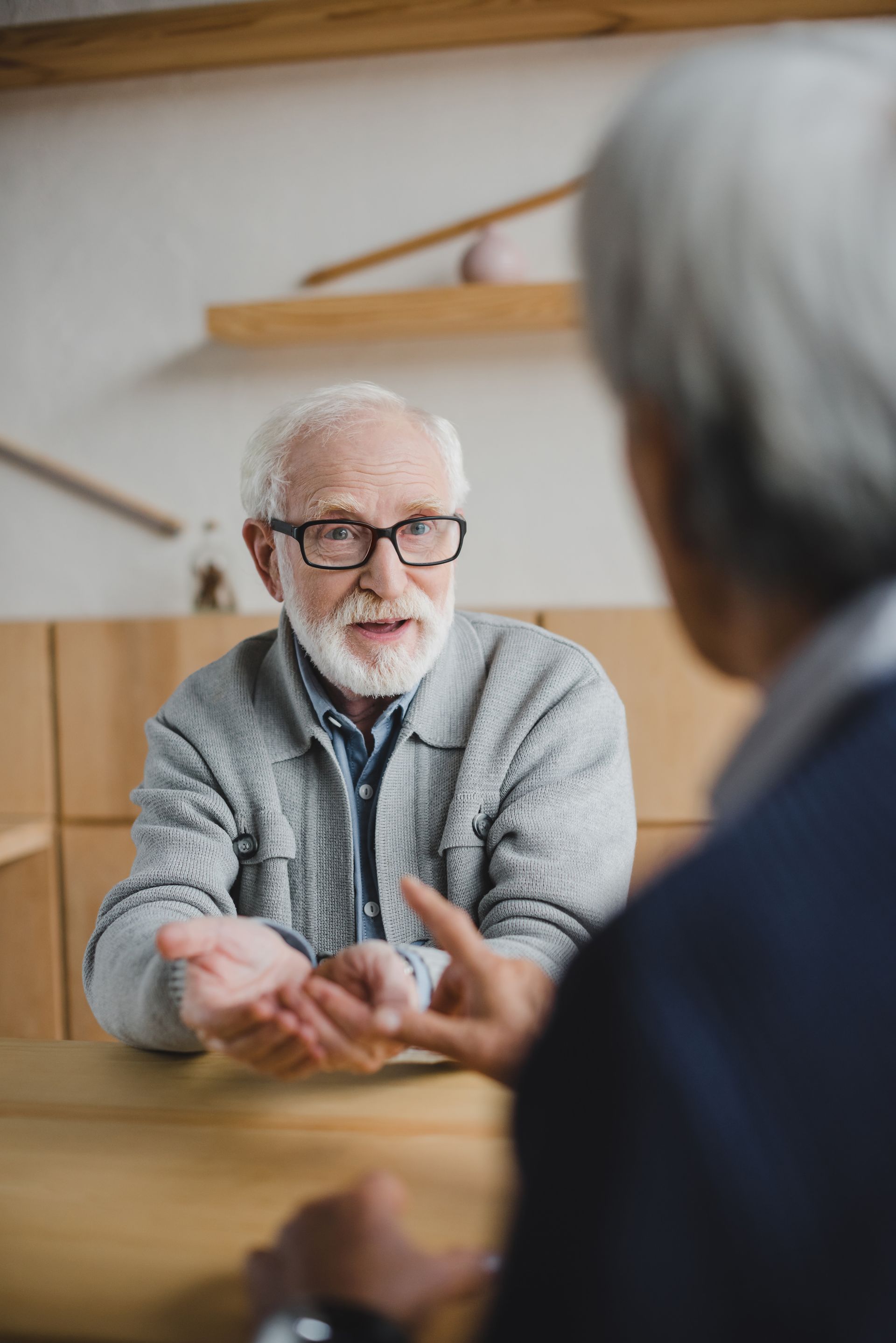 Elderly man in glasses gesturing, talking to another person at a table.