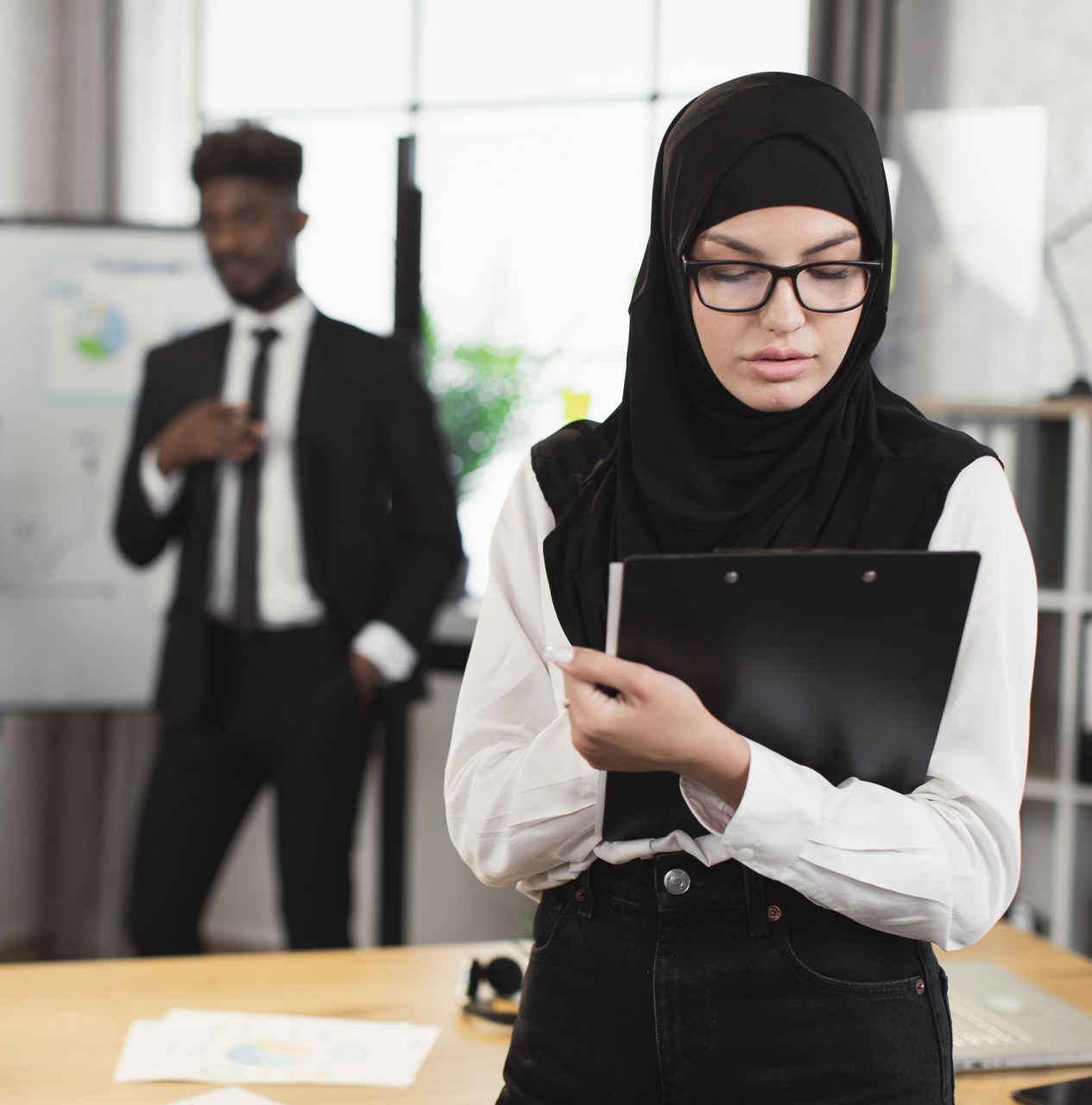 Woman in hijab and glasses, holding a clipboard, looking down in an office setting with colleagues in the background.