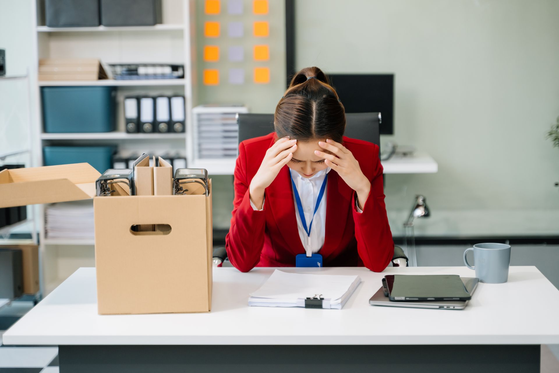 Woman in red blazer at desk, head in hands, beside box of belongings, likely laid off.