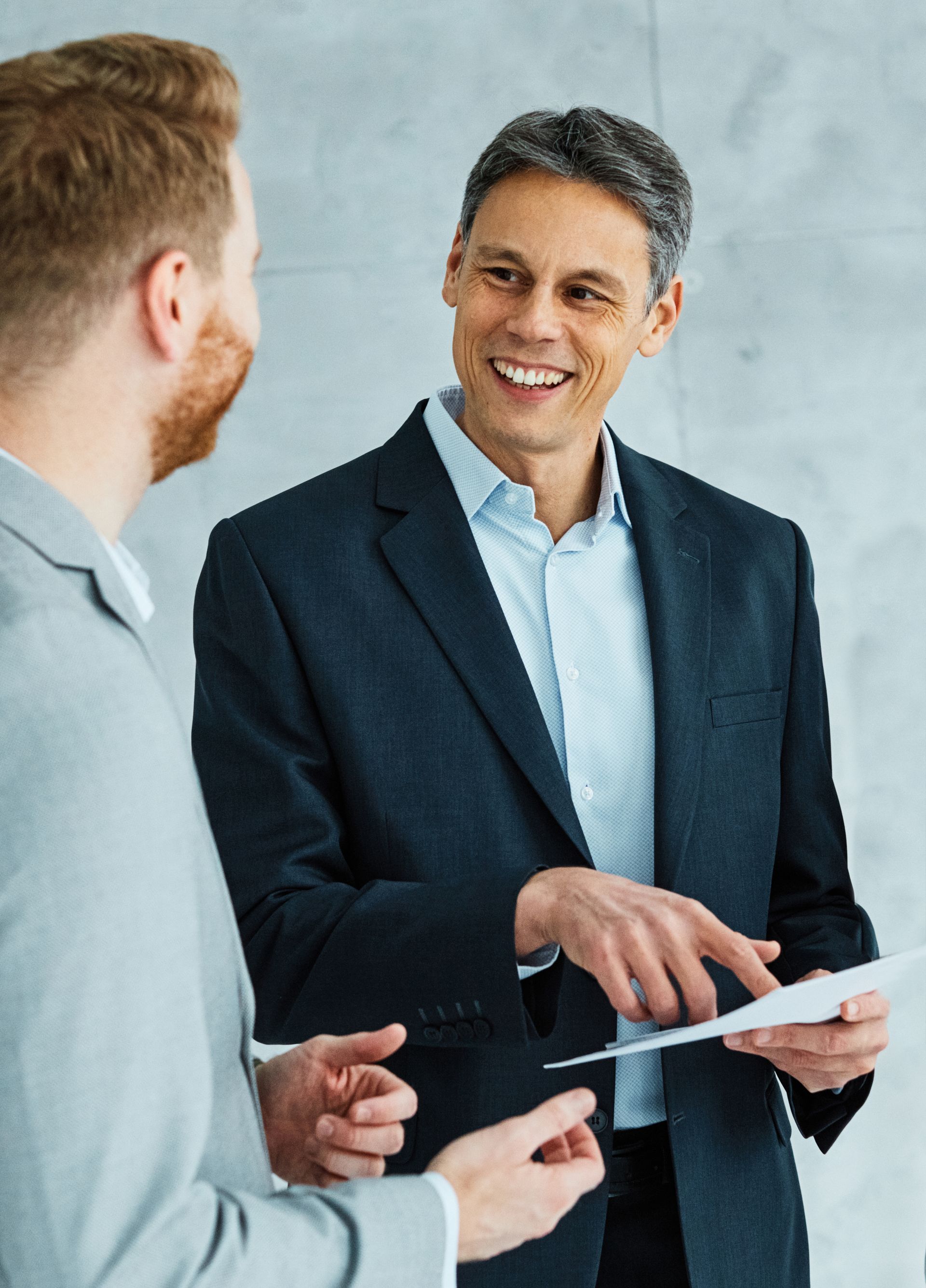Two men in suits discussing paperwork; one points, smiles. Indoor, neutral background.