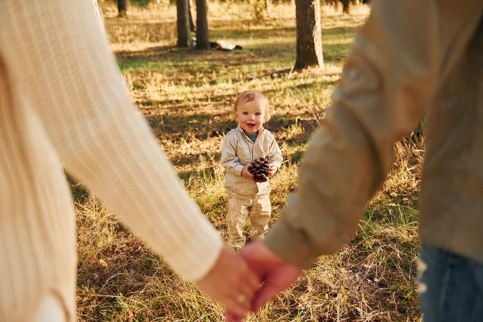 Parents holding hands, focused on a smiling toddler holding a pinecone in a sunlit forest.