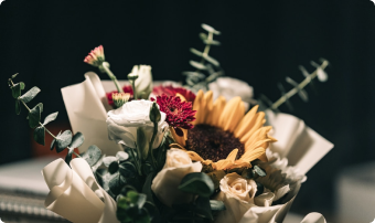 Bouquet of mixed flowers with white paper wrapping and greenery, featuring a yellow sunflower and pink blooms