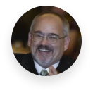 Smiling man with glasses and mustache in a dark suit and tie, seated indoors.