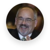 Smiling man with glasses and mustache in a dark suit and tie, seated indoors.