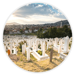 Rows of white gravestones on a hillside cemetery overlooking a city under a cloudy sky
