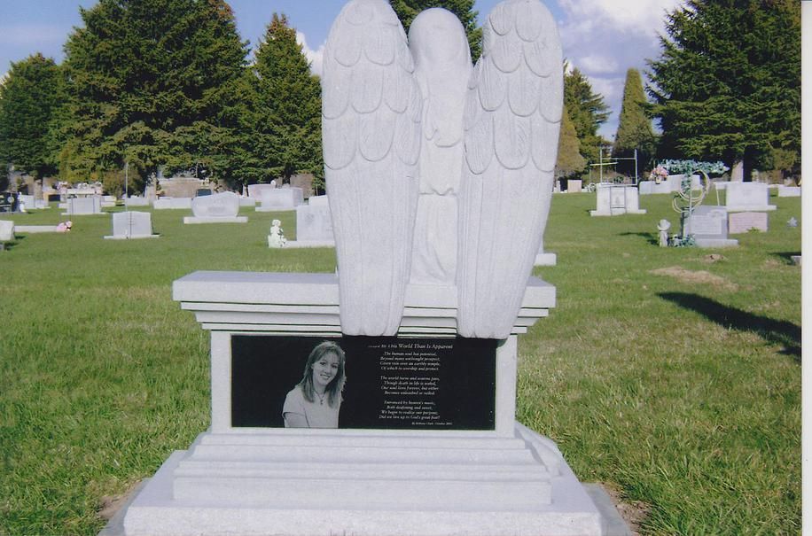 White angel gravestone monument in a cemetery with a black memorial plaque.