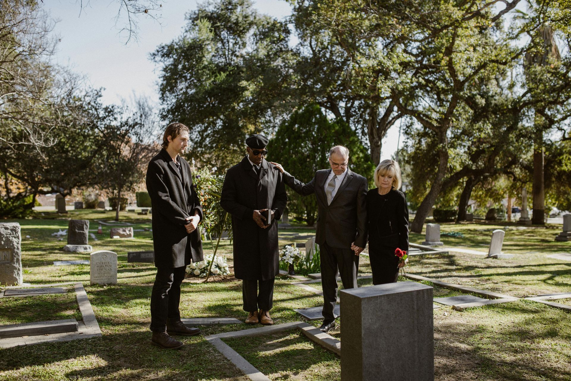 Four people stand among gravestones in a sunny cemetery, facing a headstone in a somber moment.