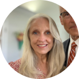 Smiling blonde woman in white top, with a man in a suit partially visible behind her.