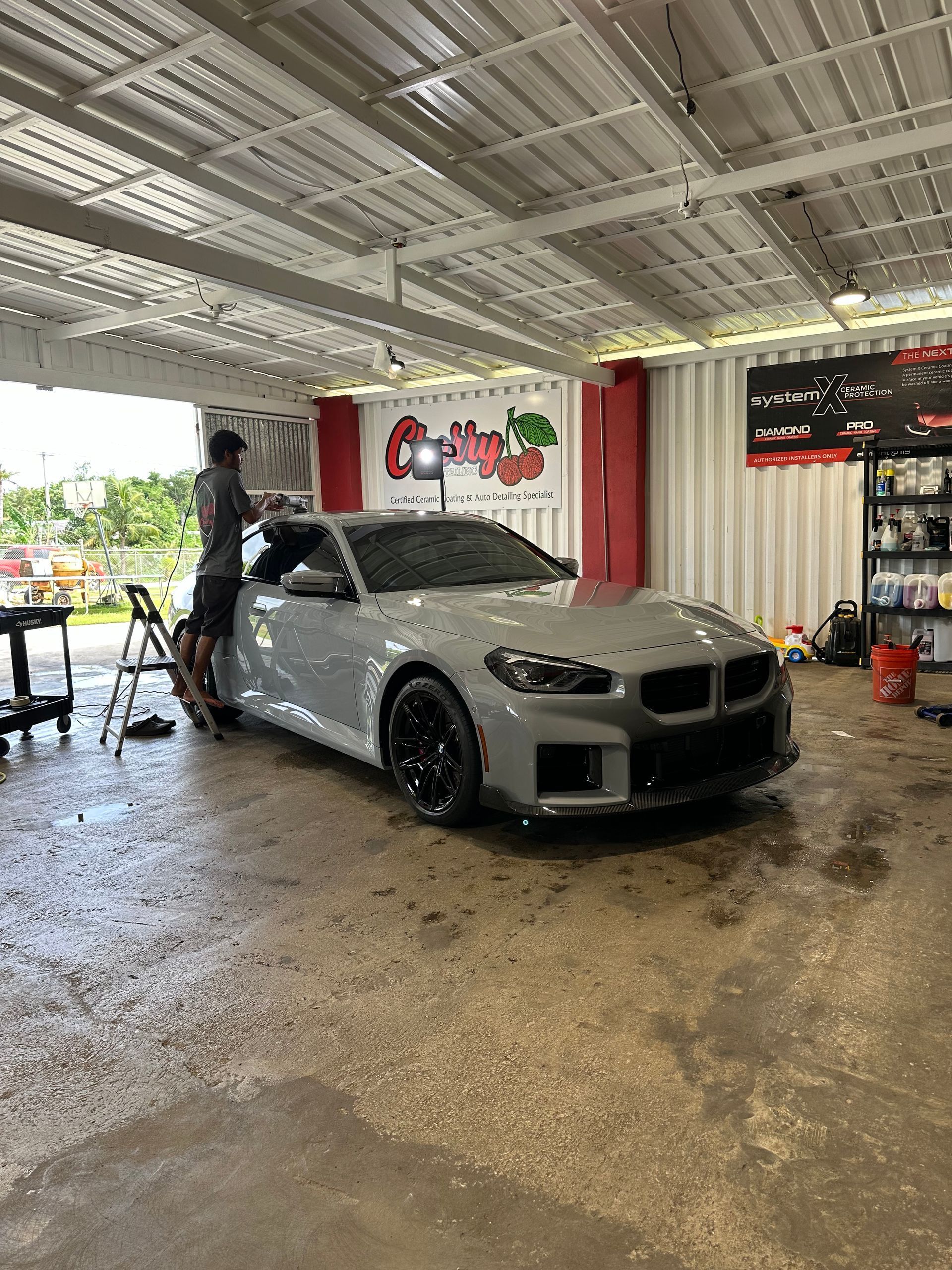 A person in a garage applies a wrap or coating to a gray BMW sports car.