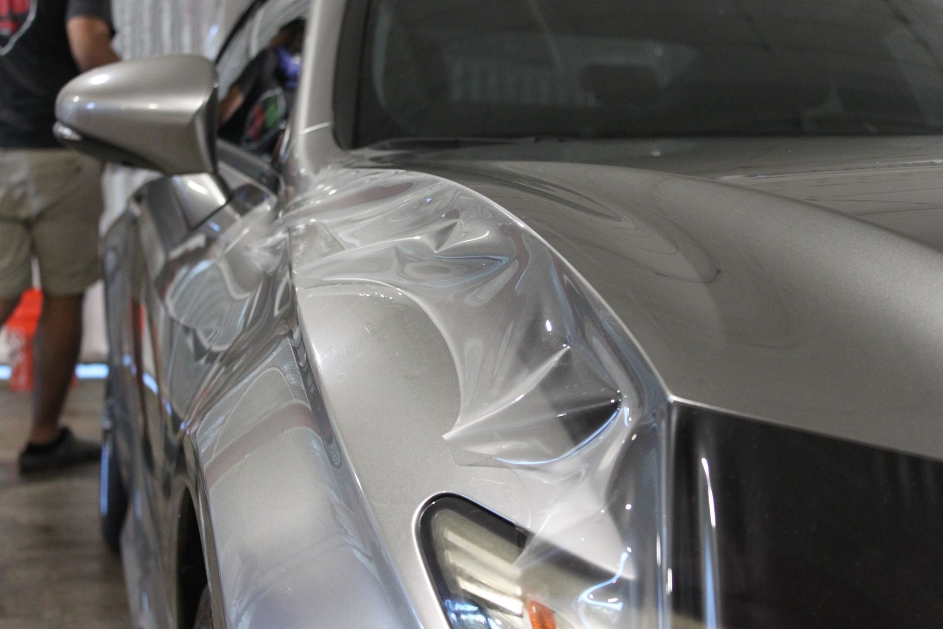 A person applying a clear protective film onto the silver fender of a car in a workshop.